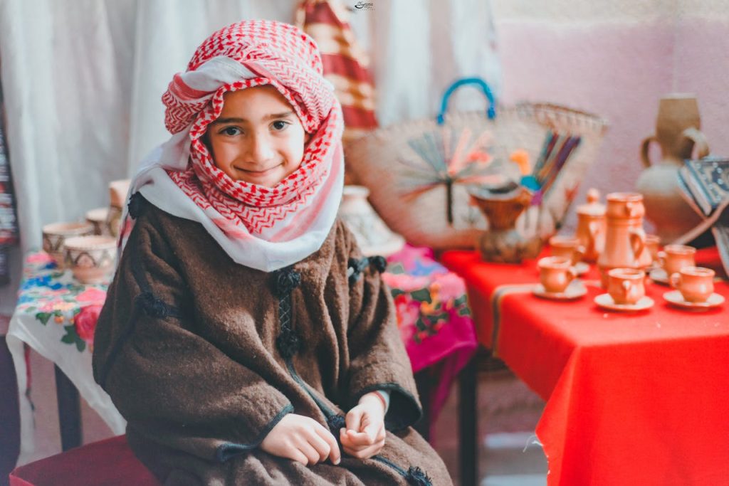 Home A cheerful child wearing a keffiyeh sitting indoors with traditional decor, showcasing cultural heritage.